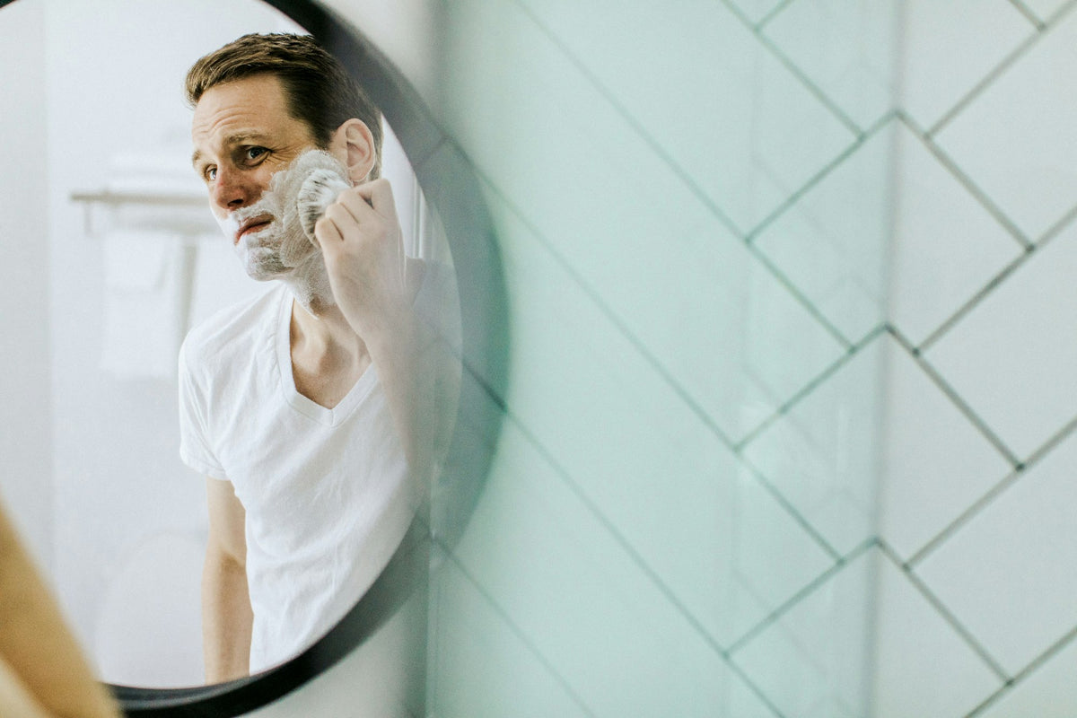 A man with shaving lather on his face shaves with a razor in front of a bathroom mirror in soft morning light