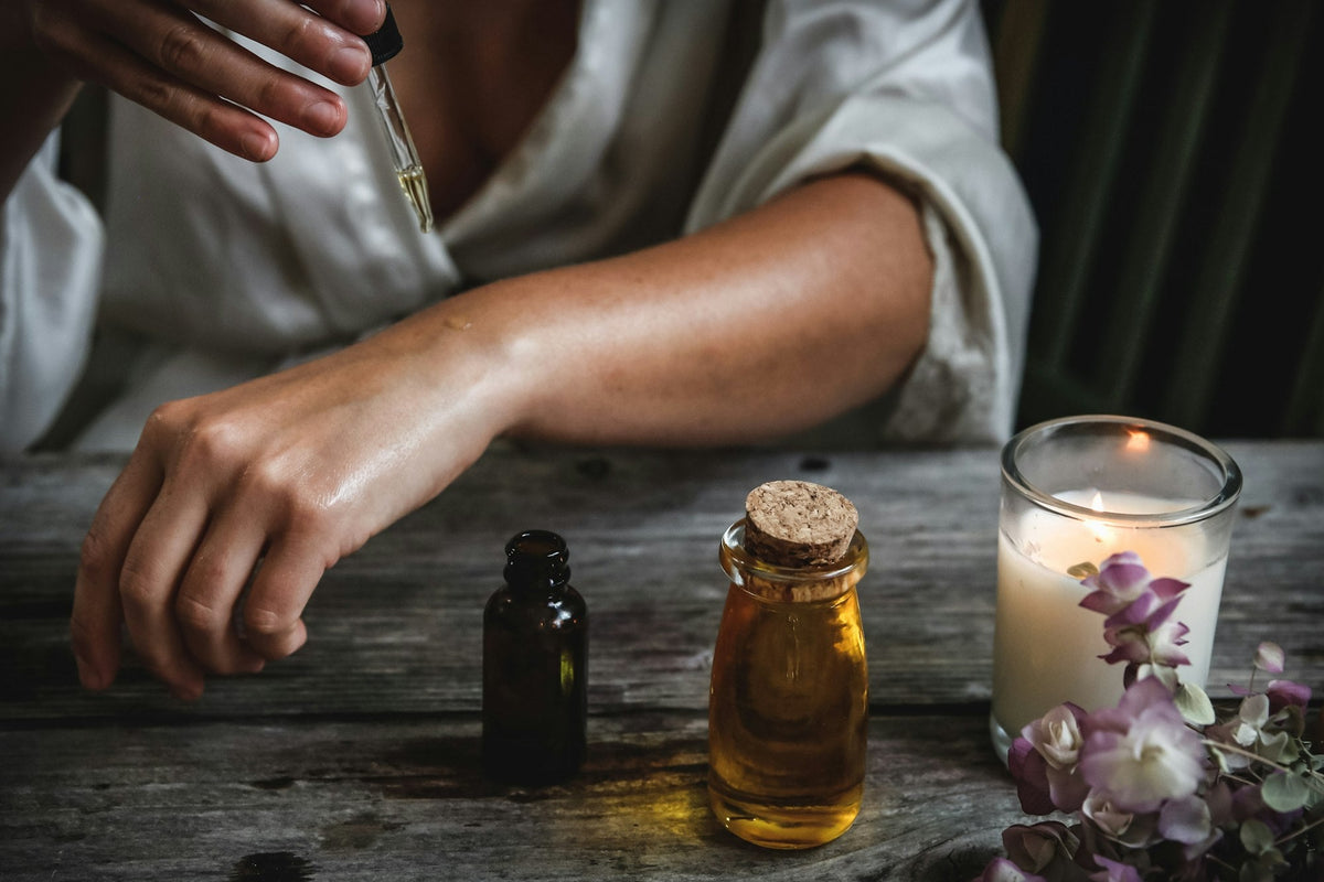 Close-up of hands applying oil from a glass dropper onto a bare arm at a sunlit wooden table