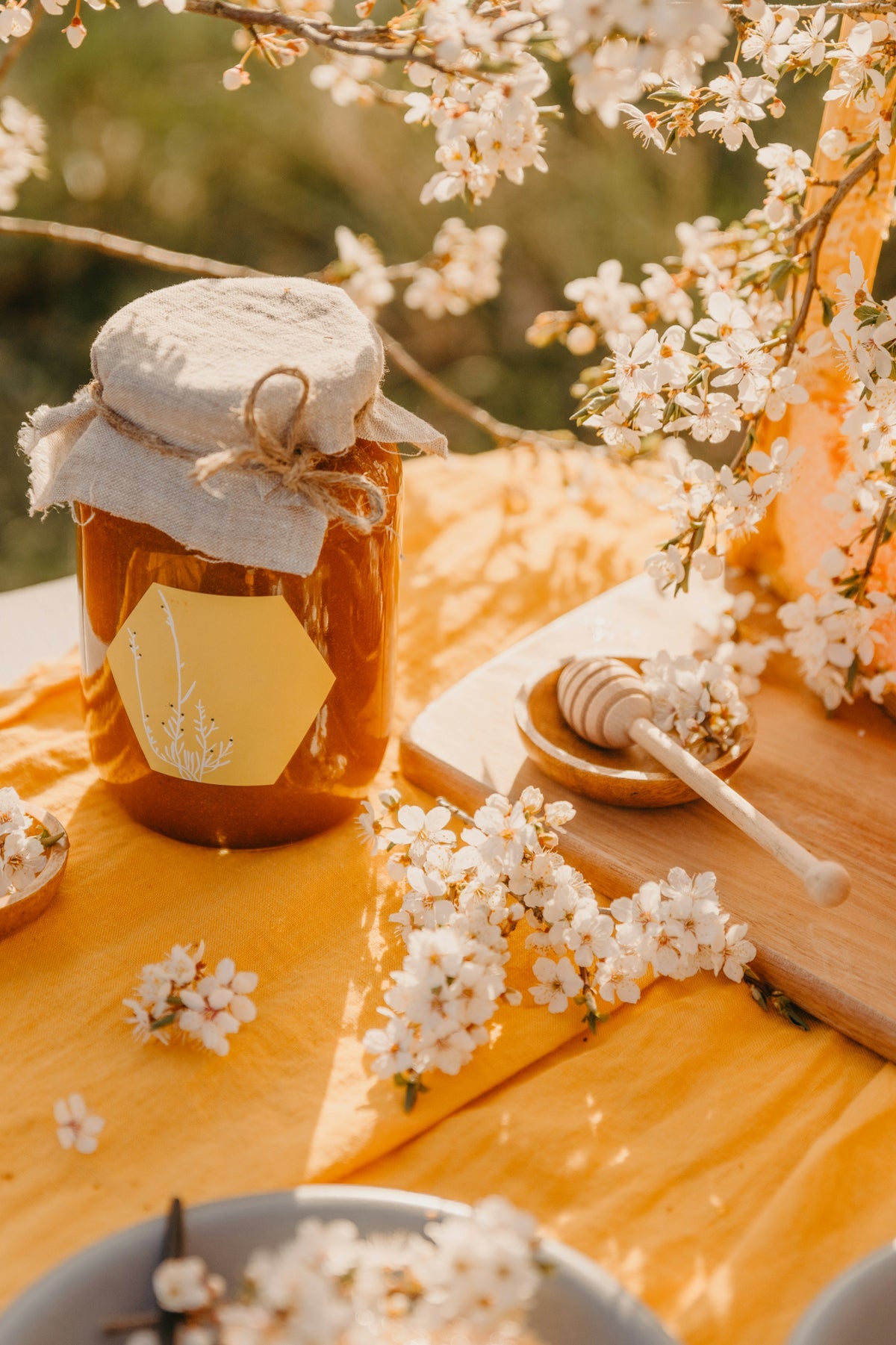 A glass jar of golden raw honey on a table beside fresh spring flowers in soft, warm natural light